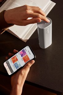 Bílý bezdrátový reproduktor Sonos Roam SL držený v ruce spolu se smartphonem zobrazujícím aplikaci Sonos, pohled shora