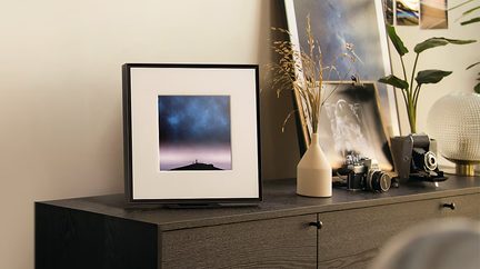 Sort kvadratisk ramme med foto af stjernehimmel på mørkt træmøbel, beige vase med tørrede græsser, vintage kameraer, kugleformet lampe