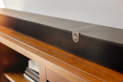 Svart ljudlåda i metallnät, sedd i snedvinkel, med texturerat frontgaller, slät övre yta med pekskärmstangenter och ett cirkulärt silverfärgat emblem med Klipsch-logotypen, placerad på en trämöbel
