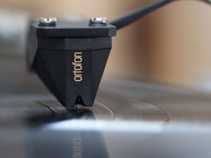 Črna mat plastična fonokapsula Ortofon, približava prednjo stran, turkizna igla v žlebovju vinilne plošče