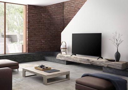 Panasonic TX-55HX940E 55 tommer fladskærms-TV, set forfra, stående på et lavt træmøbel i en moderne stue med murstensvæg