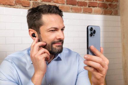 Mężczyzna w jasnej koszuli trzymający szary smartfon z potrójnym aparatem, noszący czarną słuchawkę, ściana z cegieł i białe płytki