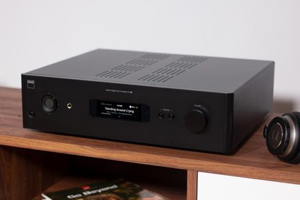 Črni hi-fi ojačevalnik NAD C 399 z osrednjim zaslonom in velikim gumbom za glasnost na lesenem regalniku