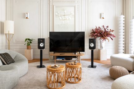 Woonkamer met flatscreen-tv, 2 luidsprekers op standaards, 2 rotan salontafels, grijze bank, vloerlamp, vaas met bloemen