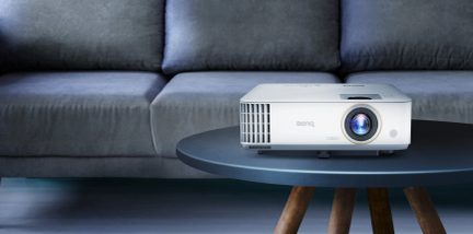 Hvid BenQ-projektor set forfra på et gråt sofabord, med en grå sofa i baggrunden