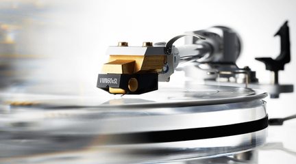 Guldfarvet metal pickup til pladespiller med sort plastikhus, set i profil i ultranærbillede, monteret på en sølvfarvet arm over en gennemsigtig vinylplade