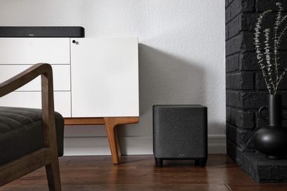 Denon Home Subwoofer svart, frontvy, placerad på golvet nära en fåtölj och en tegelkamin