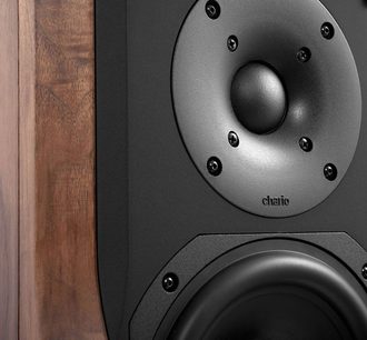 Chario Constellation Delphinus MK2, close-up frontal, caixa com acabamento em nogueira de veios visíveis, painel frontal preto fosco com tweeter de cúpula central rodeado por seis parafusos aparentes e woofer parcialmente enquadrado