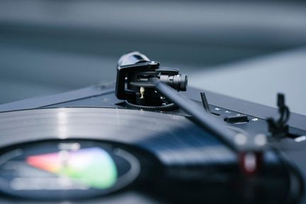 Plattenspieler in Nahaufnahme mit einem schwarzen Tonarm, der schräg auf eine schwarze Vinyl-Schallplatte zeigt, mit einem mehrfarbigen Etikett in der Mitte.