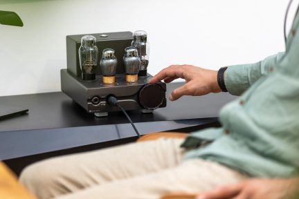 Černý elektronkový audio zesilovač s velkým ovladačem hlasitosti, položený na stole, ruka při nastavování