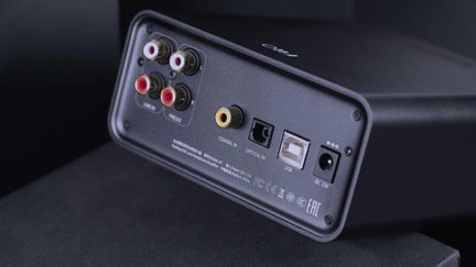 Matte black rectangular aluminum audio case, three-quarter rear view, featuring six red and white RCA connectors, a yellow coaxial input, a square optical port, a USB-B port, and a 12V power socket