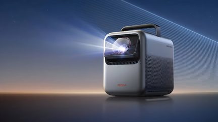 Metallic gray portable projector with an aluminum casing, front lens framed in black emitting a light beam, black top handle, perforated side grille, right front three-quarter view