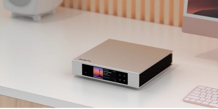Caja de audio hi-fi rectangular plateada de aluminio cepillado, pantalla a color y botones táctiles en el panel frontal, aletas de ventilación negras en el lado derecho, vista en tres cuartos desde el frente derecho