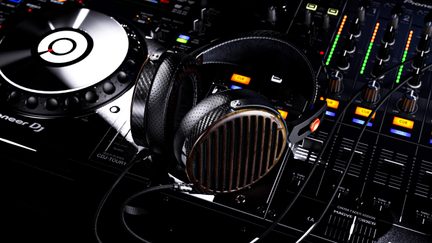 Over-ear hovedtelefoner i sort læder med kornet struktur og carbonfiber, ørepuder med kobberfarvet gitter og aftageligt kabel, set i tæt opstilling fra oven i skrå vinkel placeret diagonalt på en DJ-konsol
