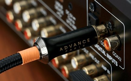 Černý audio koaxiální kabel s opleteným pláštěm, černý lesklý kovový válcový konektor s pozlaceným nápisem „ADVANCE PARIS“ a rýhovaným stříbrným kroužkem, detailní boční pohled zapojený do RCA konektoru s oranžovou vložkou na zadním panelu hi-fi zařízení