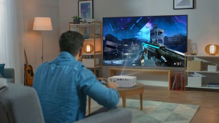 Moderní obývací pokoj, osoba zezadu s ovladačem, velký černý televizor zobrazující hru, dřevěný konferenční stolek a bílý projektor