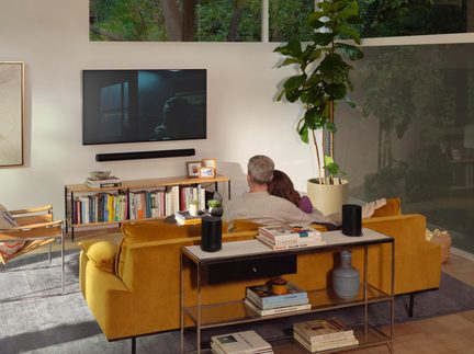 Moderne woonkamer, achteraanzicht van een koppel op een mosterdgele stoffen bank, wand-tv, soundbar, boekenkast en plant
