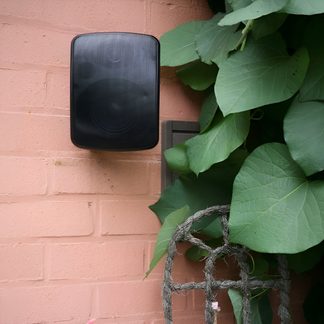 Cassa acustica nera da esterno fissata su un muro in mattoni rosa accanto a un fogliame verde e fitto