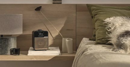 Musta digitaaliradio, jossa on näyttö ja puiset lamelliristikot, edestä kuvattuna puisen yöpöydän hyllyllä olevien kirjojen päällä.