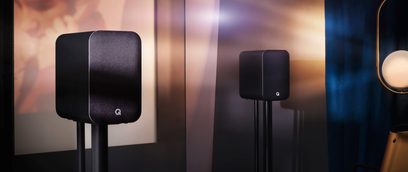 To sorte Q Acoustics M20 højttalere set forfra på en nedtonet kobber- og mørkblå baggrund