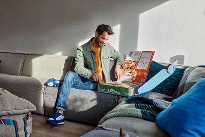 Mand siddende på en grå sofa, læsende i farverige brochurer, iført blå sneakers og en grøn og orange sweater