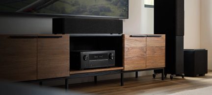 Amplificateur audio-vidéo noir à façade métallique avec grand écran numérique central et deux boutons rotatifs latéraux, vue de face dans une niche ouverte d’un meuble TV en bois