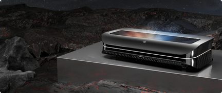 Sort og sølvfarvet laserprojektor i aluminium, lavt rektangulært kabinet med afrundede hjørner, frontgitter og stort glasparti på toppen, set i trekvart forfra-perspektiv
