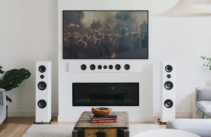 Woonkamer, muurtelevisie boven een open haard, twee witte kolomluidsprekers, centraal kanaal, houten salontafel