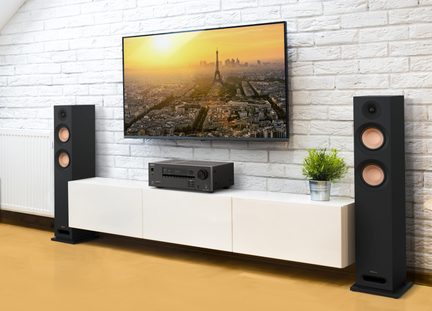 Frontansicht einer Heim-Audio-Video-Anlage mit schwarzem, wandmontiertem Flachbildfernseher, zwei schwarzen Standlautsprechern mit doppelten kupferfarbenen Lautsprechern und Bassreflexöffnung unten, sowie einem schwarzen Verstärker auf einem weiß lackierten Wandmöbel