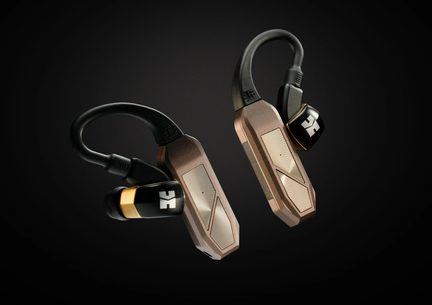 To trådløse in-ear-ørestik med rektangulær stang i kobberrosa metal, fleksibel sort ørekrog og sort cylindrisk øreprop med guldring, vist i skrå vinkel på mørk baggrund