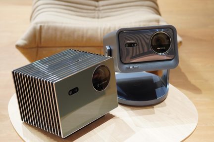 Två moderna videoprojektorer, den ena rektangulär med spegelyta i metallgrå färg och kromade sidokanter, den andra i antracitgrå med stort objektiv i ring monterat på en svängbar bas, presenterade i ett lätt överhängande perspektiv på ett rundbord i ljust trä.