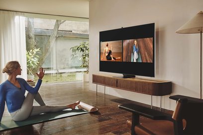 Kvinde, der gestikulerer mod et Samsung QE75QN800B 2022 TV, der viser en video, set fra siden i en moderne stue