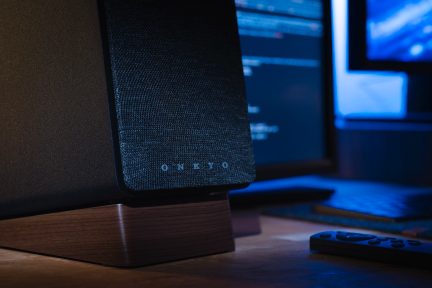 Zwarte luidspreker met stoffen rooster, houten basis, zwarte afstandsbediening, computerscherm en toetsenbord op de achtergrond