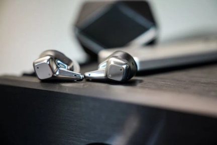 Kaldvaates lähivõte kahest hõbedasest, läikiva metallviimistlusega juhtmevabast kõrvasisesest kõrvaklapist, millest kummalgi on lühike nurgeline vars, asetatud tumedale pinnale