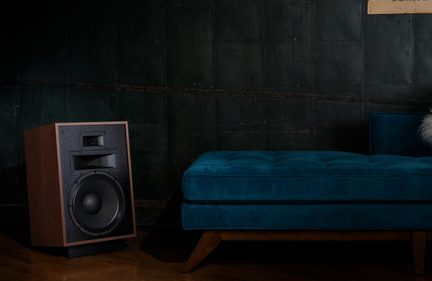 Hi-Fi reproduktor z dřeva zobrazený ze tří čtvrtin, s černým texturovaným čelem, pavilonem a velkým wooferem, umístěný vedle modré pohovky