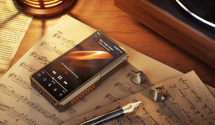 Reproductor de audio Hi-Fi FiiO dorado de metal, con la pantalla encendida y auriculares intrauditivos apoyados sobre partituras y escritorio de madera