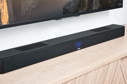 Černý soundbar Canton umístěný pod televizorem na světlém dřevěném TV stolku, čelní panel s modrým LED displejem