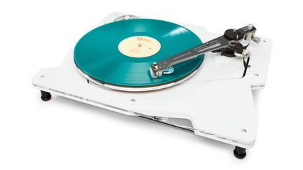 Platenspeler van bovenaf gezien, licht schuin, met een glanzend witte acryl basis en transparante randen, een ronde draaitafel met een turquoise transparante vinylplaat met een crèmekleurig label, een zwarte toonarm met metalen elementen, zwarte dempende voetjes