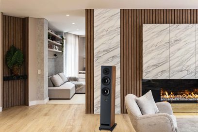Gulvhøyttaler Davis Acoustics Krypton 6 Classik sett forfra, med 3 svarte høyttalerelementer, rektangulær sokkel, plassert på et lyst tregulv, omgitt av en vegg i marmor og tre, lysegrå lenestol, peis med flammer, hyller og beige sofa i bakgrunnen.