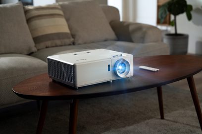 Optoma UHZ50 hvid og sort videoprojektor, set forfra på et lavt, mørkt træbord foran en grå sofa i stuen