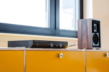 Čierny hi-fi zosilňovač Musical Fidelity A1 na žltom nábytku vedľa hnedého reproduktora s čiernou prednou stranou