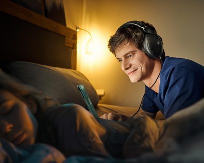 Persona che indossa delle cuffie, sdraiata su un letto, che tiene in mano un tablet, con una lampada accesa sullo sfondo