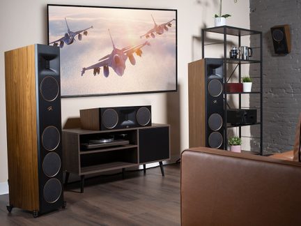 To store højttalere i træ, et møbel med centerhøjttaler, en metalhylde, en brun sofa, en skærm med fly