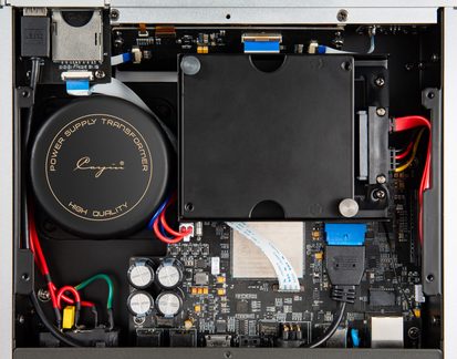 Top view of the inside of a silver metal electronic device enclosure, featuring a black toroidal transformer with gold silkscreen, a rectangular black drive enclosure, a black circuit board fitted with silver cylindrical capacitors, and red, black, green, and blue wiring
