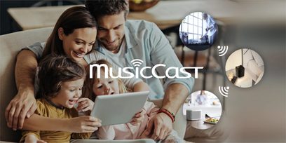 Billedet viser en smilende familie på fire personer, der sidder sammen på en sofa og ser på en tablet-skærm, med ikoner relateret til musik og trådløs forbindelse omkring dem