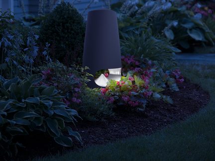 Mat zwarte cilindrische tuinlamp van metaal met gebogen uitsnede die licht naar de grond schijnt, frontaal gezien tussen bloeiende planten