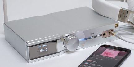 Stříbrný obdélníkový kryt s digitálním displejem, kruhovým tlačítkem, audio konektory, bílými sluchátky a černým smartphonem