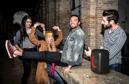 Imaginea prezintă un grup de patru tineri adulți zâmbitori și energici care pozează împreună pe o stradă pavată în fața unei clădiri vechi din cărămidă, cu expresii fericite și prietenoase