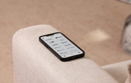 Black smartphone in a matte plastic case, screen on, seen from a slight three-quarter overhead angle, placed on the armrest of a textured beige fabric sofa