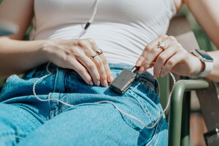 Primo piano delle mani con anelli, custodia audio portatile nera metallizzata con clip e cavo intrecciato argentato su jeans blu